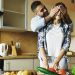 Young man covering girlfriends eyes with hands and surprising her in the kitchen at home. Loving couple cooking breakfast in the morning